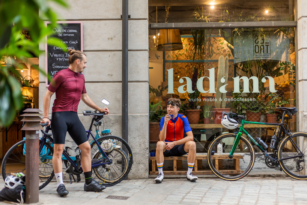 2 Maenner in Radtrikots mit Fahrraedern vor einem Cafe. Das Radtrikot ist bordeaux Rot und das andere Blau orange.