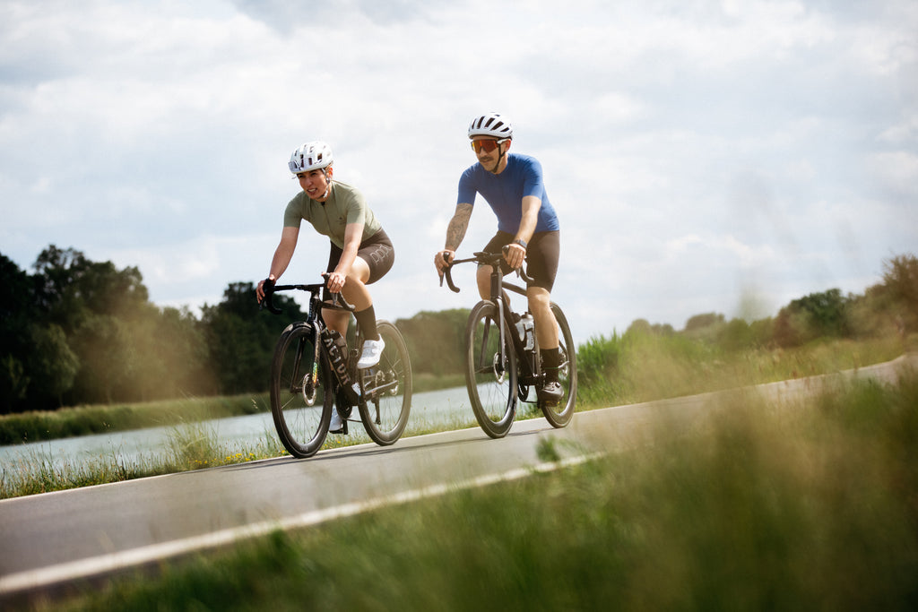 Zwei Radfahrer aus der Seitenansicht – Mann im blauen und Frau im grünen Rennradtrikot auf der Strasse.