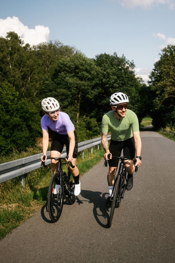 Frontalaufnahme von zwei Rennradfahrern – Frau im lila Trikot und Mann im grünen Trikot auf der Straße unterwegs