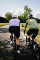 Rückansicht von zwei Rennradfahrern – Frau im lila Trikot und Mann im grünen Trikot auf der Straße unterwegs