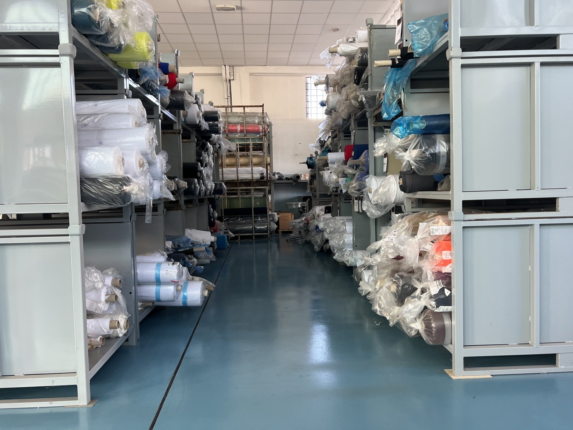 Lagerraum mit Regalen voller Stoffrollen und Textilien, ordentlich verpackt in Folie, in einem hellen Produktions- oder Materiallager für Rennradbekleidung.